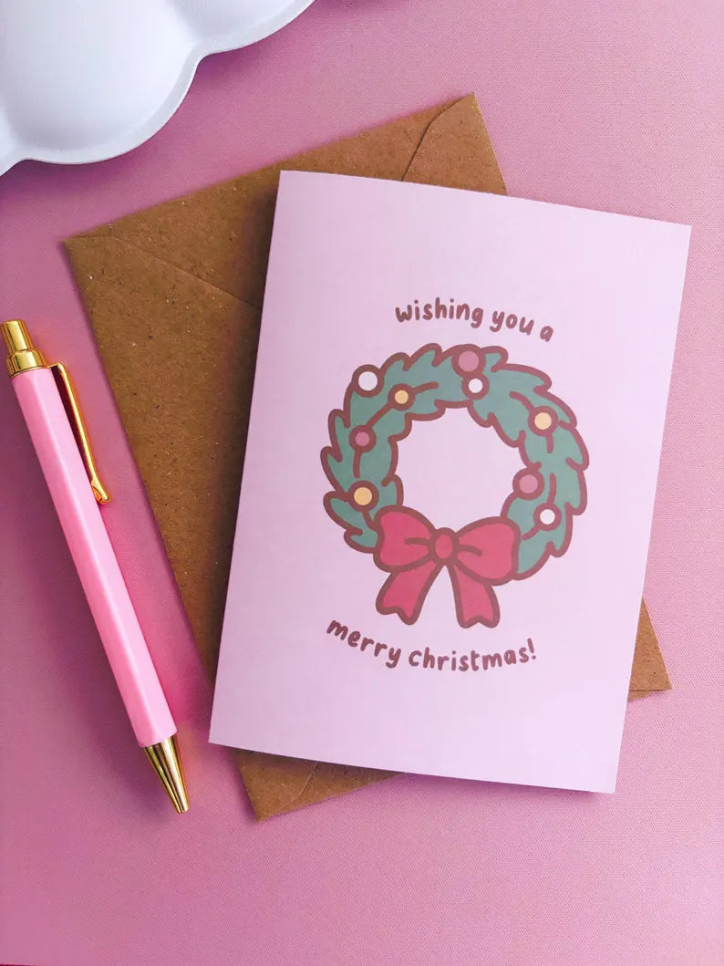 Christmas card with wreath and bow design, pink pen, and brown envelope on pink background