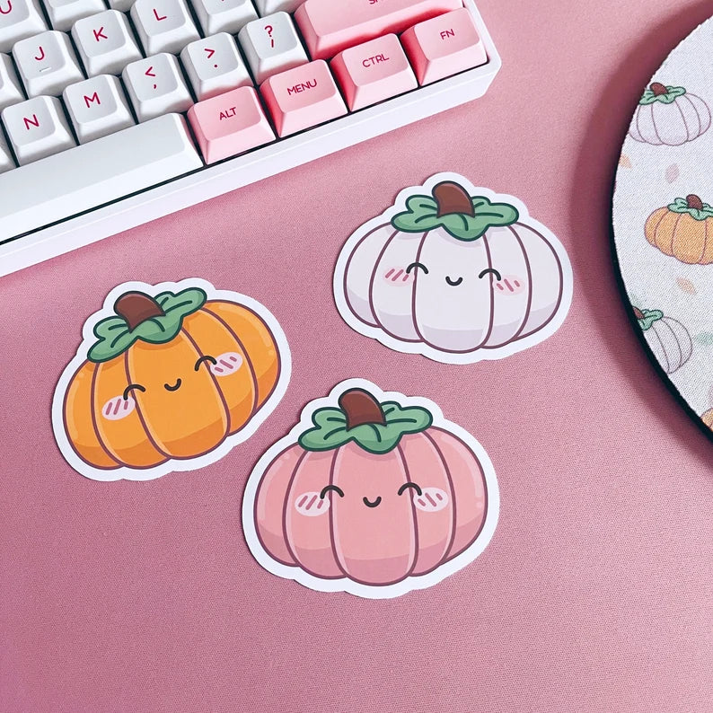 Three cute pumpkin stickers on a pink surface with a keyboard in the background.
