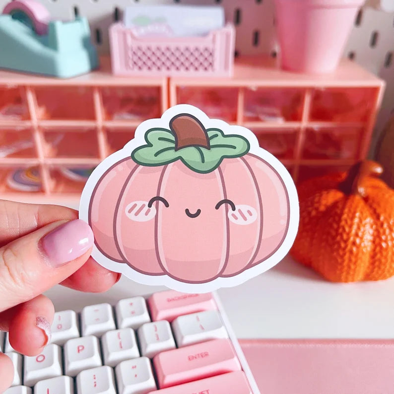 Hand holding a cute pink pumpkin sticker with a background of a pink desk setup.