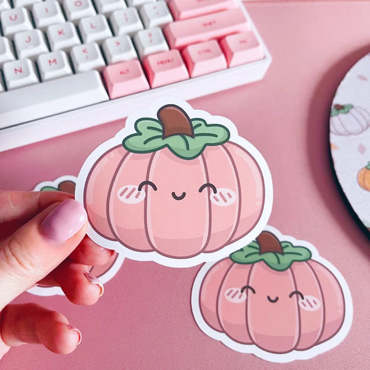 Hand holding a pink pumpkin sticker with a keyboard and mouse pad in the background