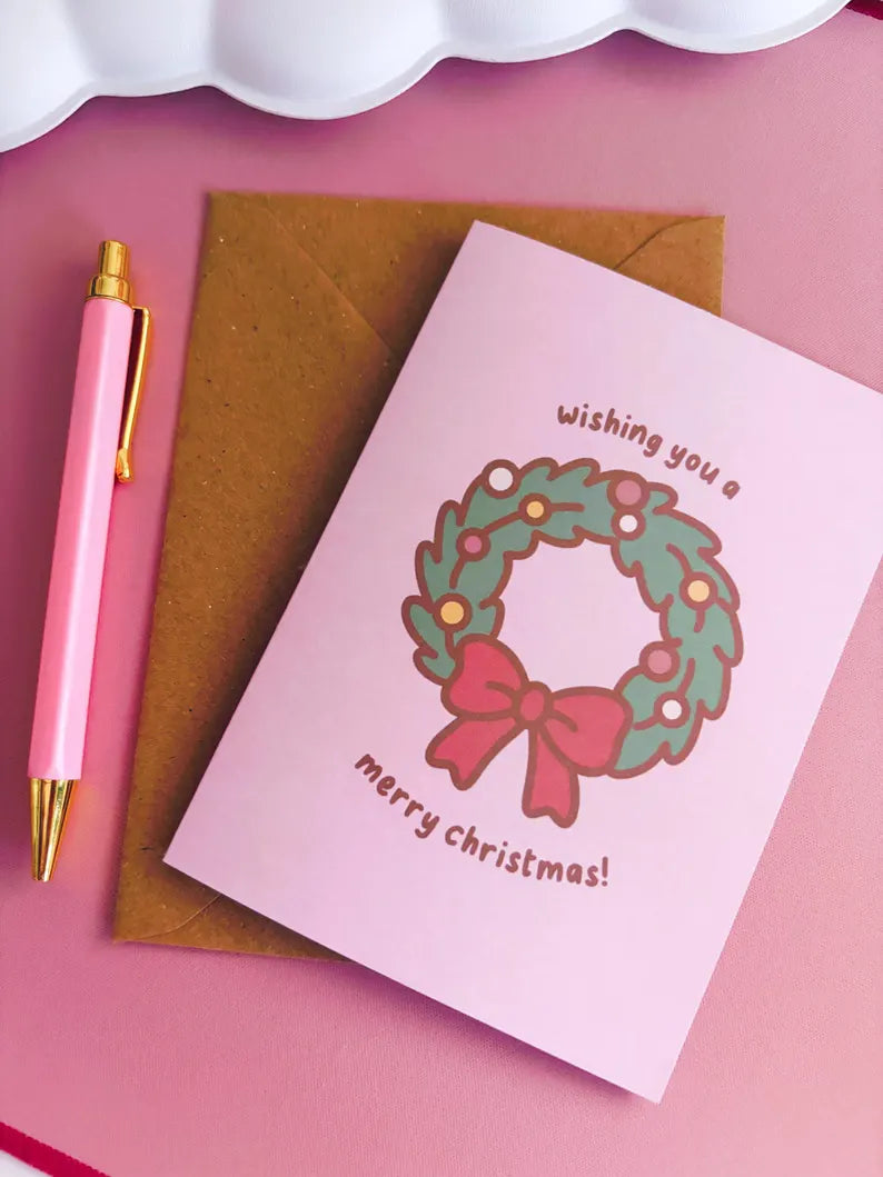 Pink Christmas card with wreath and bow design, brown envelope, and pink pen on pink background