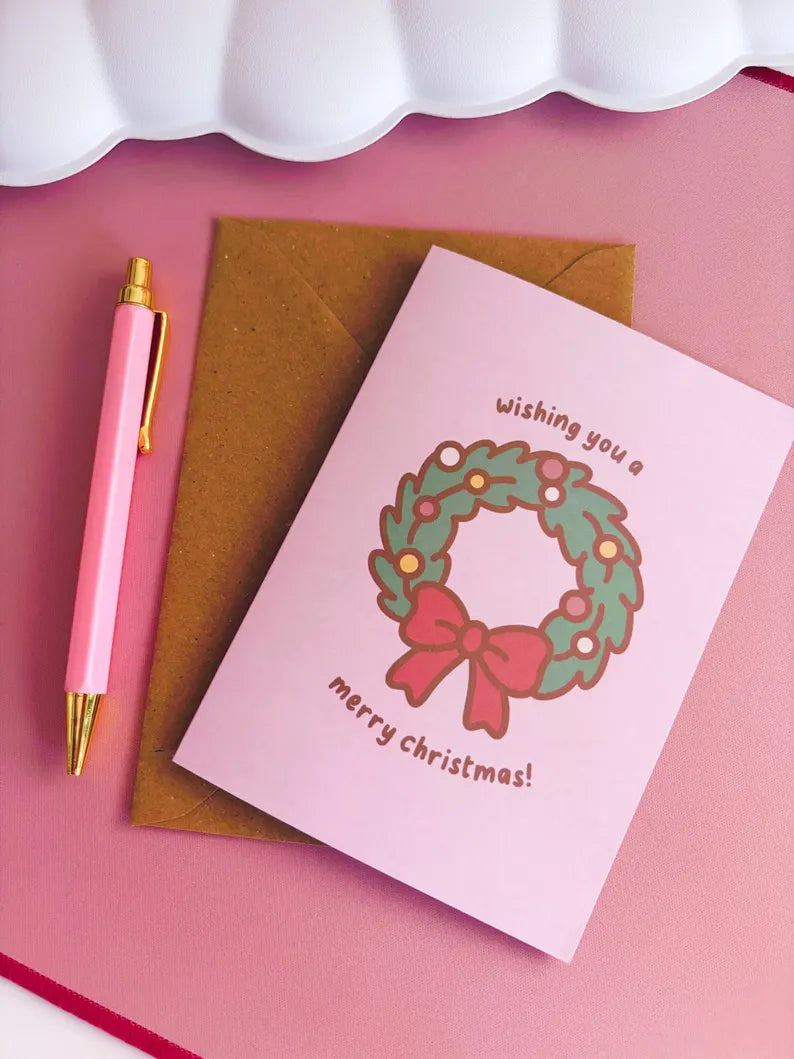 Christmas card with wreath and bow design, pen, and envelope on pink background
