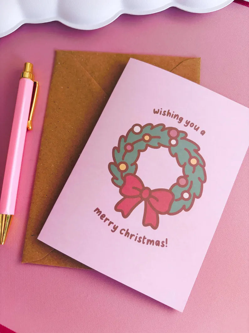 Pink Christmas card with wreath and bow design, brown envelope, and pink pen on pink background